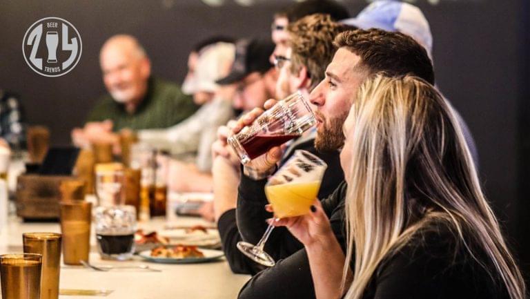 Customers drink beer at a crowded table together. SevenFifty Daily's 2024 Trends logo stamp is in the upper left corner.