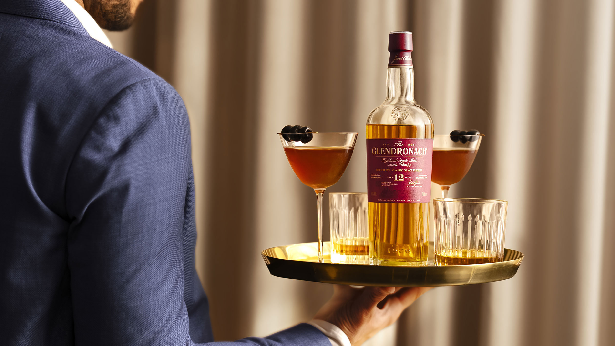 A bottle of The GlenDronach along with two cocktails are held aloft on a tray by a server